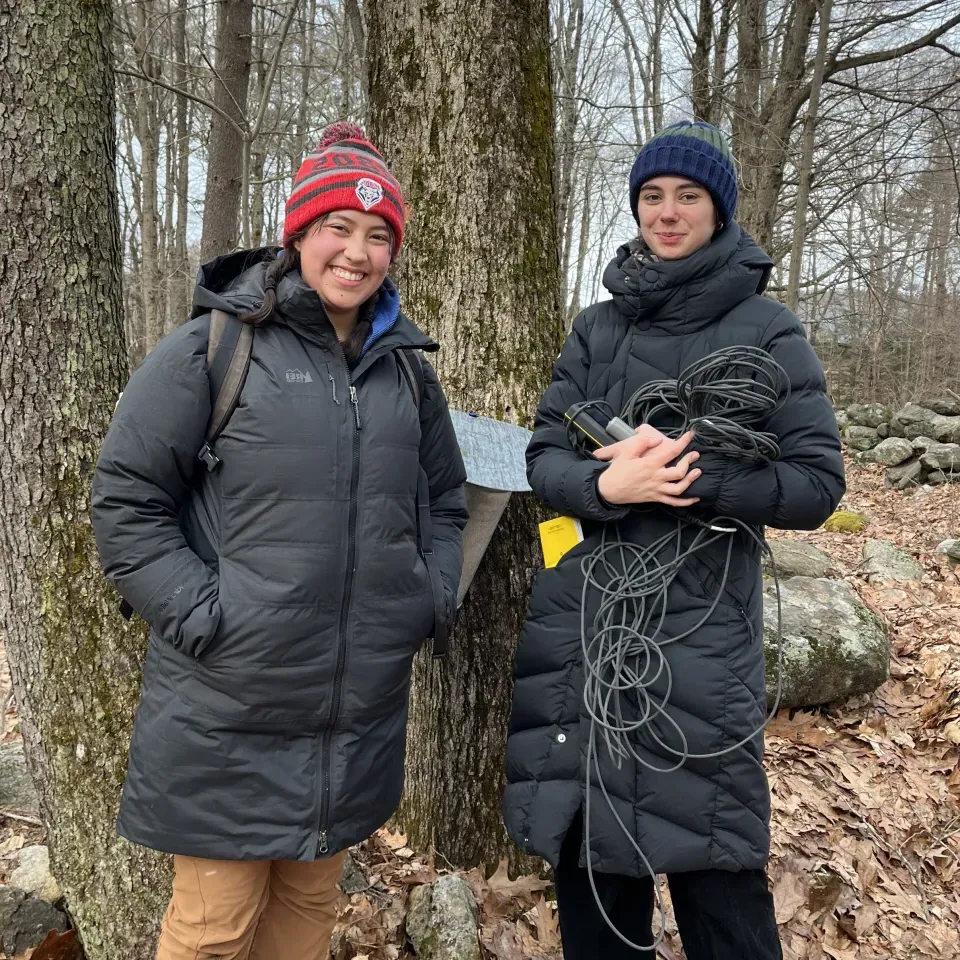 两个穿着冬衣微笑着站在树前的学生, 被落叶包围, 手持设备